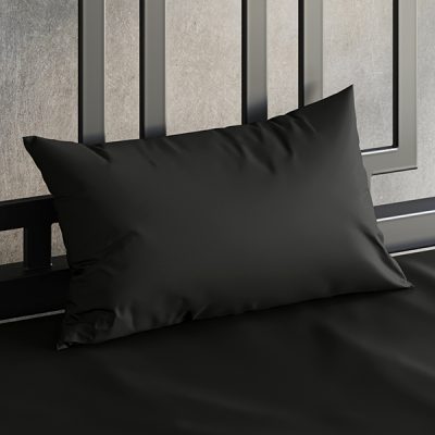 Close up of a Sheets of San Francisco Black waterproof pillow case on a bed covered in a Black Sheets of San Francisco fluidproof fitted sheet and against a black metal bedhead with the polished concrete wall behind showing through.